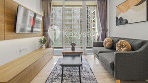 Una sala moderna con un televisor de pantalla plana, un sofá gris, un mueble de madera, una mesita con un florero y un ventanal grande con cortinas.