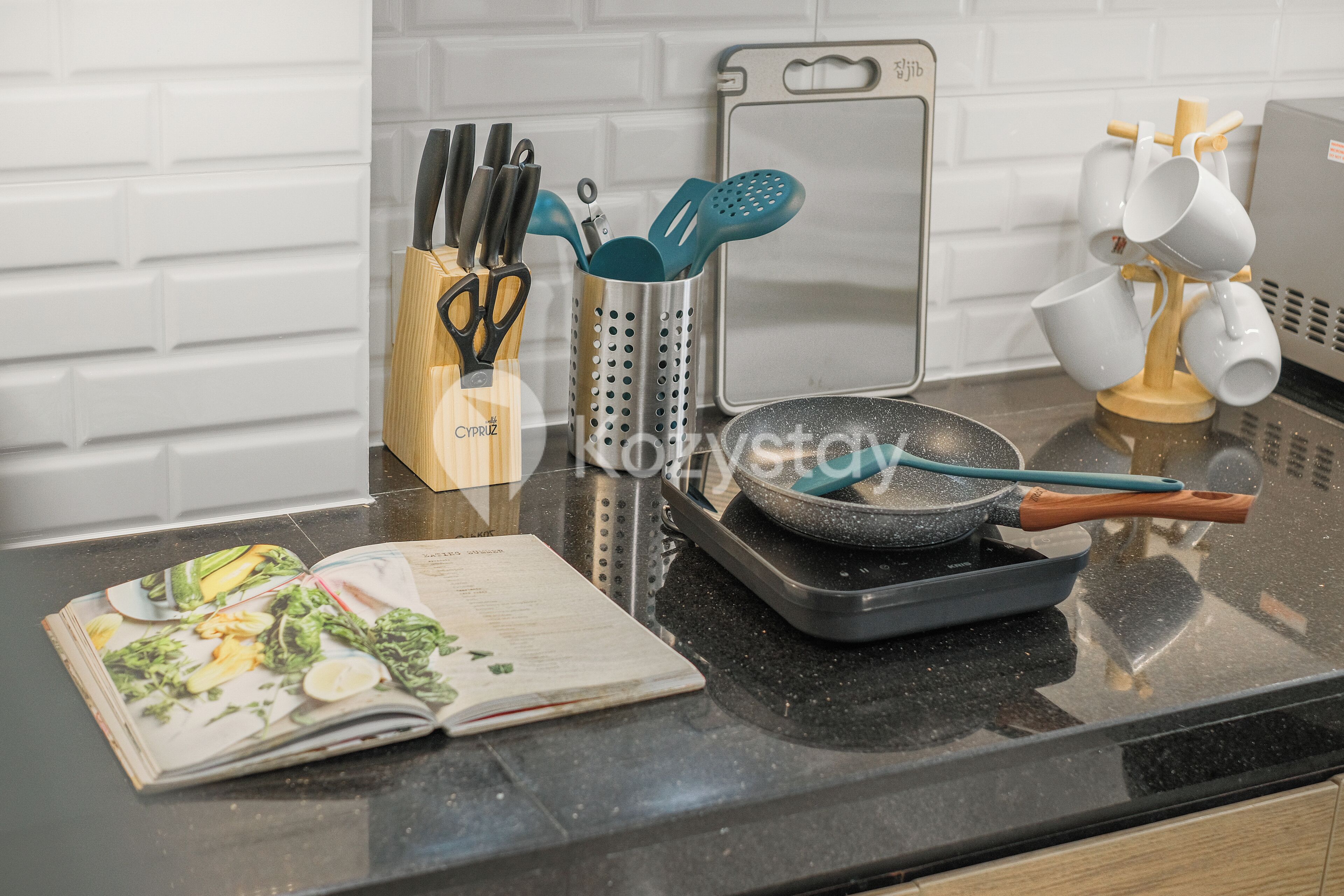 Un plan de travail de cuisine avec un livre de recettes, une poêle, un ensemble de couteaux, une râpe et un porte-tasses.