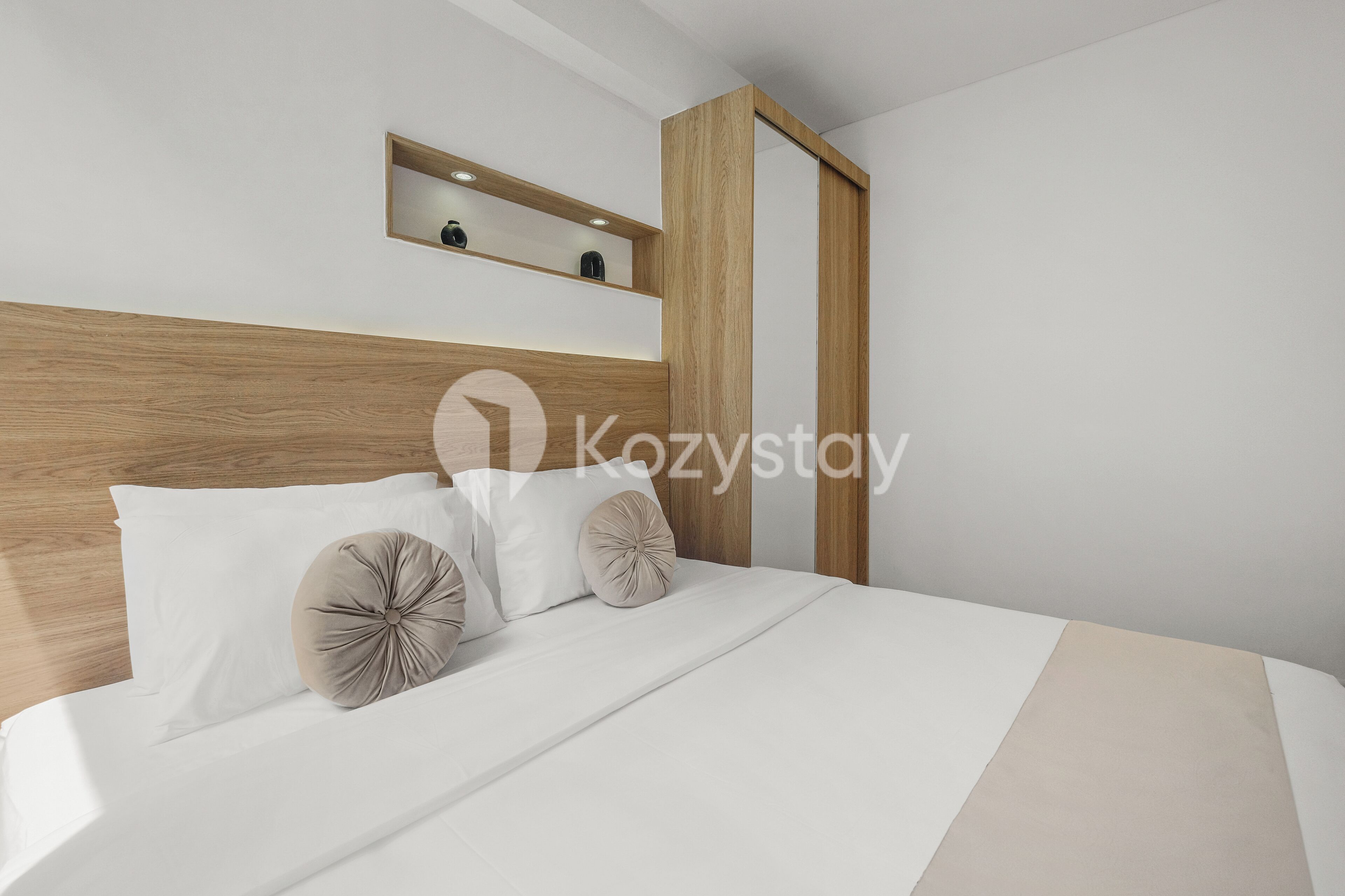 Ett modernt hotellrum med en sänggavel i trä, en säng med vita sängkläder, två dekorativa kuddar och en liten hylla ovanför sängen.