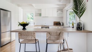 Una cocina moderna con armarios blancos, una isla central con dos sillas de mimbre, un refrigerador y un ventanal con vistas a zonas verdes.