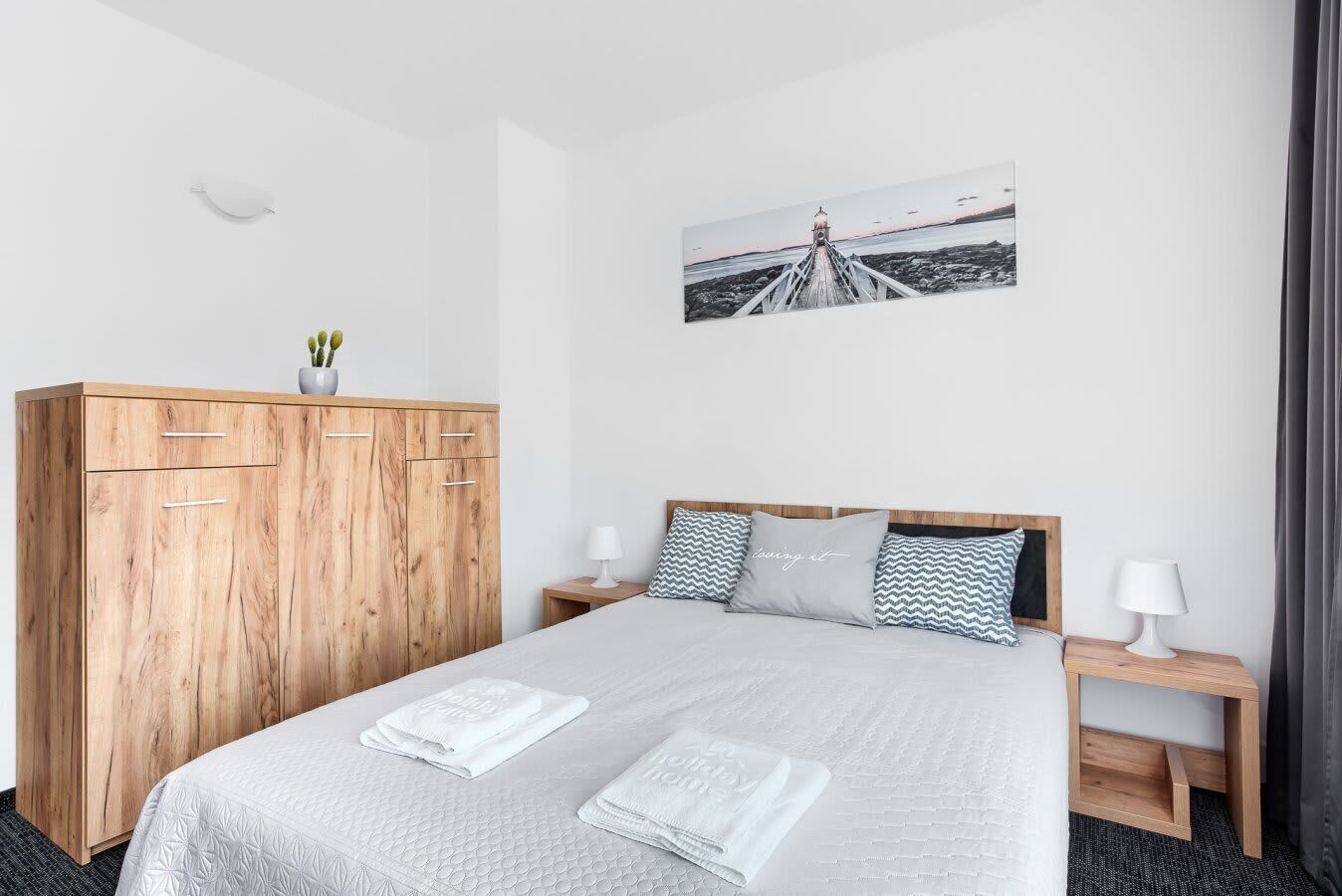 A neatly arranged bedroom featuring a wooden bed frame, folded towels at the foot of the bed, and a wall-mounted photograph above the headboard.