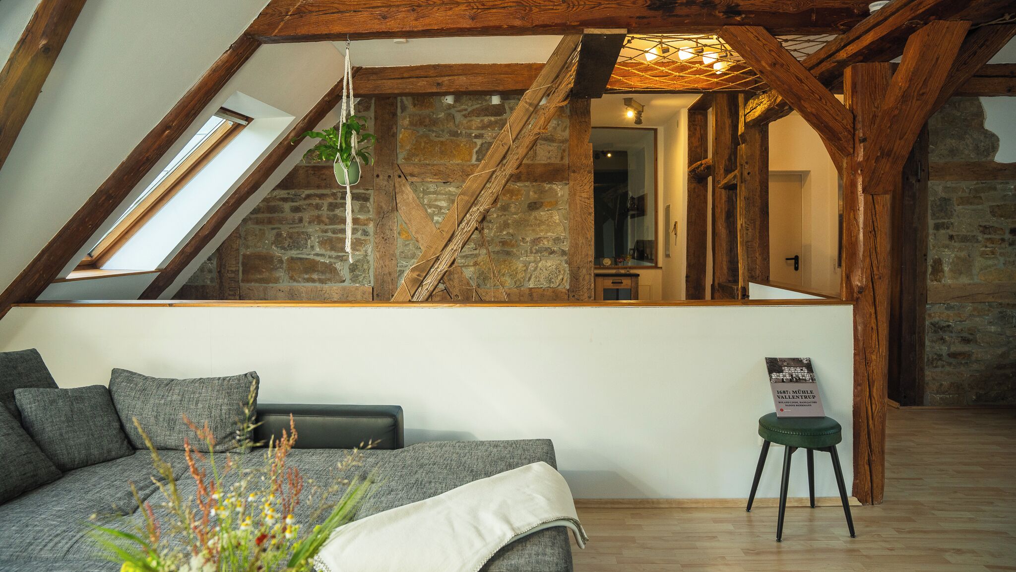 A loft-style room with a sloped ceiling, exposed wooden beams, a stone wall, a staircase, a sofa, a small table, and a hanging plant.