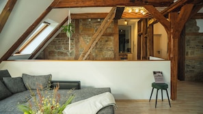 Ein Loft-Zimmer mit geneigter Decke, freiliegenden Holzbalken, einer Steinwand, einer Treppe, einer Couch, einem kleinen Tisch und einer hängenden Pflanze.