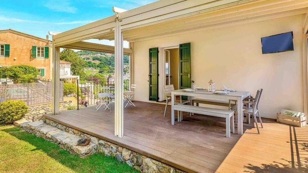 Eine überdachte Terrasse mit Esstisch und Stühlen, eine Steinmauer und Blick auf einen Hügel.