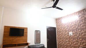 Une pièce dotée d’un ventilateur de plafond, d’un téléviseur fixé au mur, d’un papier peint à motifs, d’une porte et d’un lit recouvert d’une couverture colorée.