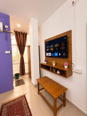 A modern living room with a mounted TV, wooden shelves, a bench, and a view of the outdoors. - The Kashi Palace (Varanasi)