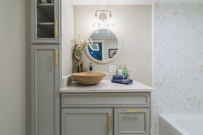 A bathroom with a wooden bowl sink, a round mirror, and a light fixture above it. There are toiletries on the counter and a towel hanging on the wall. - The Craftsman - Handcrafted escape on Pond 2BR 2BA (Norfolk)