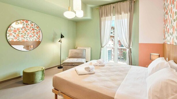 A hotel room with a bed, a round mirror, a green ottoman, and a view of buildings through the window. - Welcome to Bellini13 in La Spezia, our new holiday home recently renovated and ready to welcome you for an unforgettable stay! The interiors have been renovated with pastel colors that create a relaxing and serene atmosphere and with wallpaper that adds  (La Spezia)