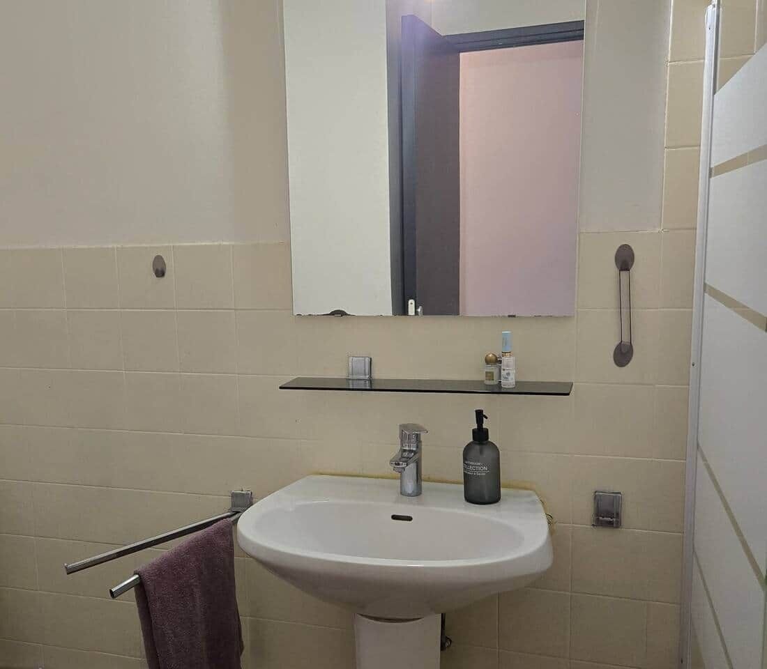 Une salle de bain avec un lavabo blanc, un miroir au-dessus, un porte-serviettes et une étagère contenant des articles de toilette.