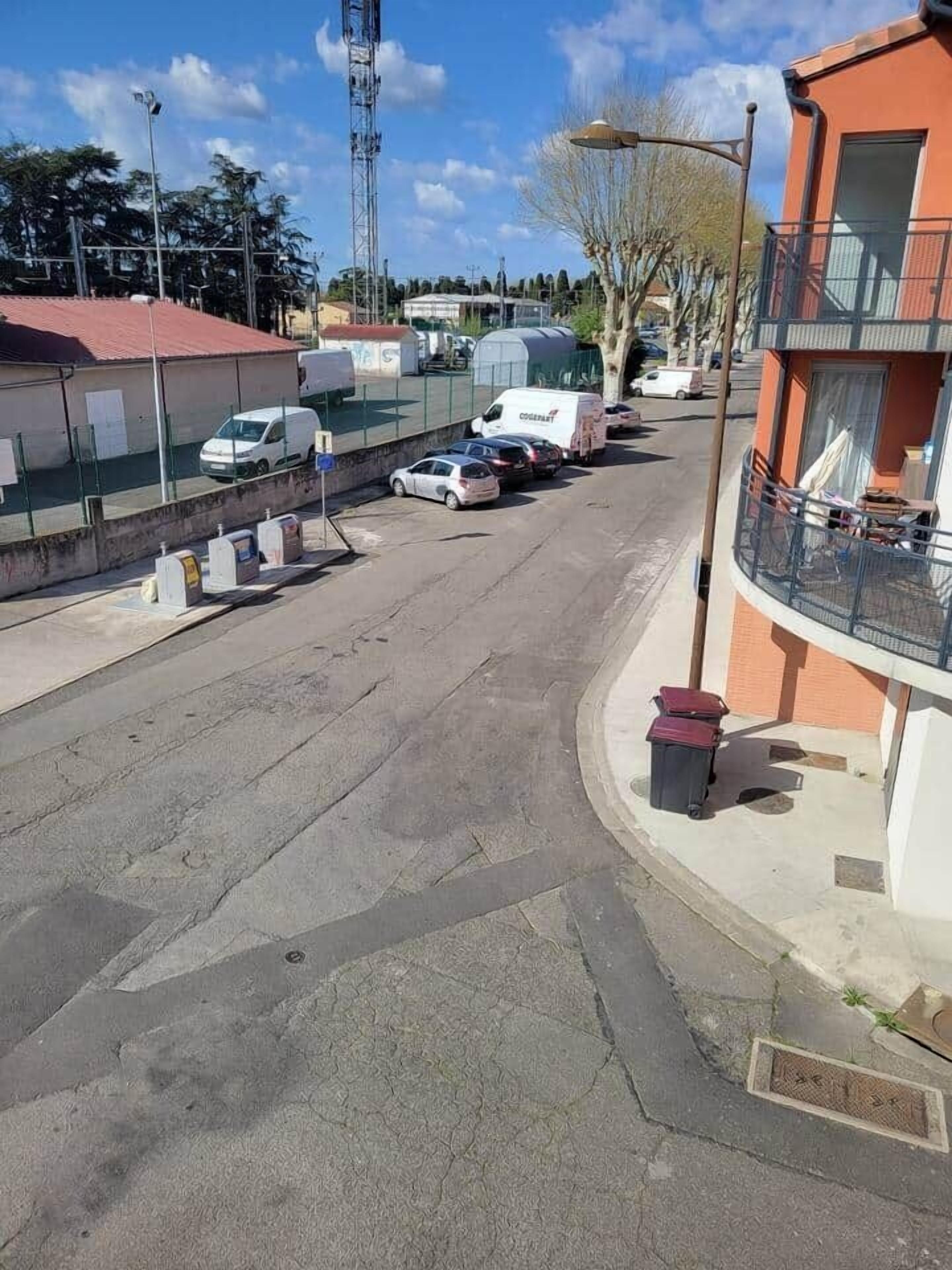 A street view with parked cars, a building with a balcony, and a clear sky.