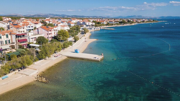 An aerial view of a coastal town with a clear beach, a pier extending into the sea, and residential buildings with red roofs. - Apartment Adriatic With Pool (Vodice)