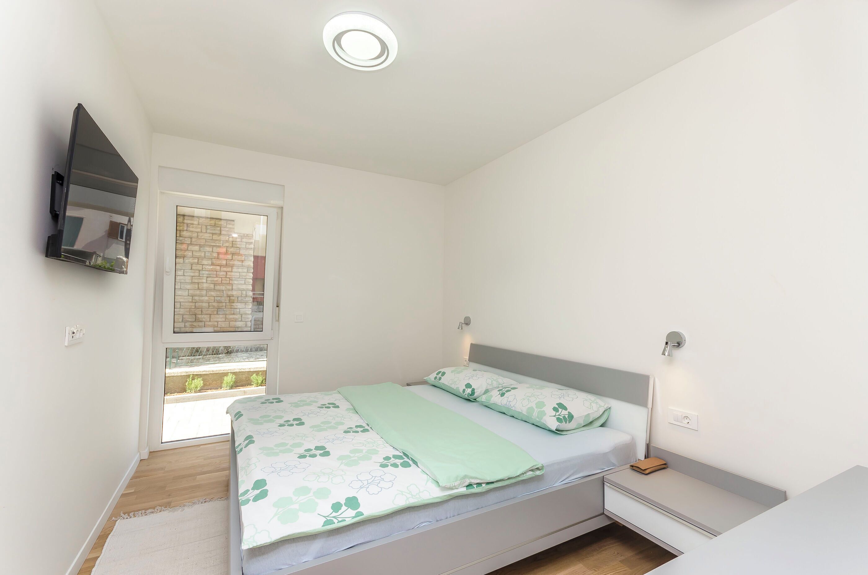 Un dormitorio con cama, mesitas de noche, televisor de pantalla plana montado en la pared y ventana con vista a un edificio de ladrillo.