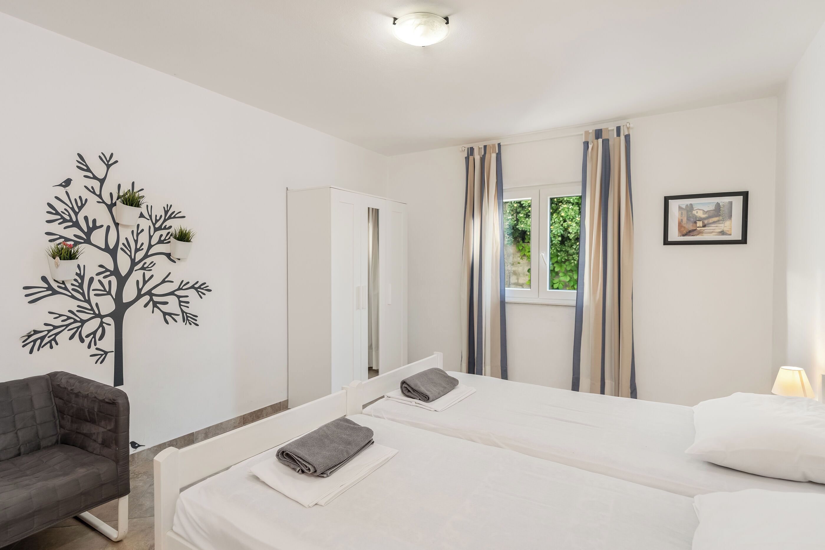 A bedroom with two beds, a chair, a window with curtains, and a wall decoration.