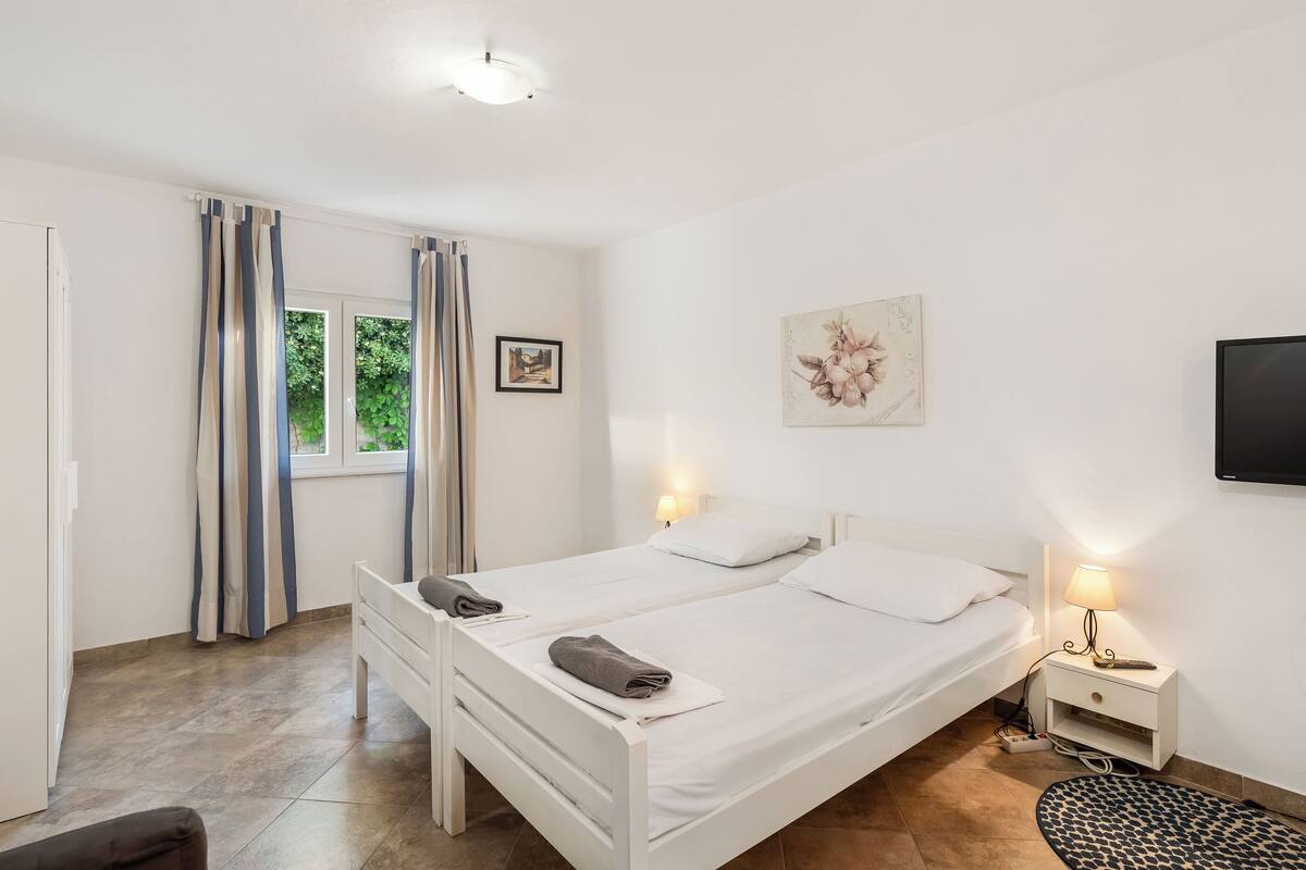 Une chambre avec deux lits, une table de chevet avec une lampe, une fenêtre avec des rideaux et un téléviseur fixé au mur.