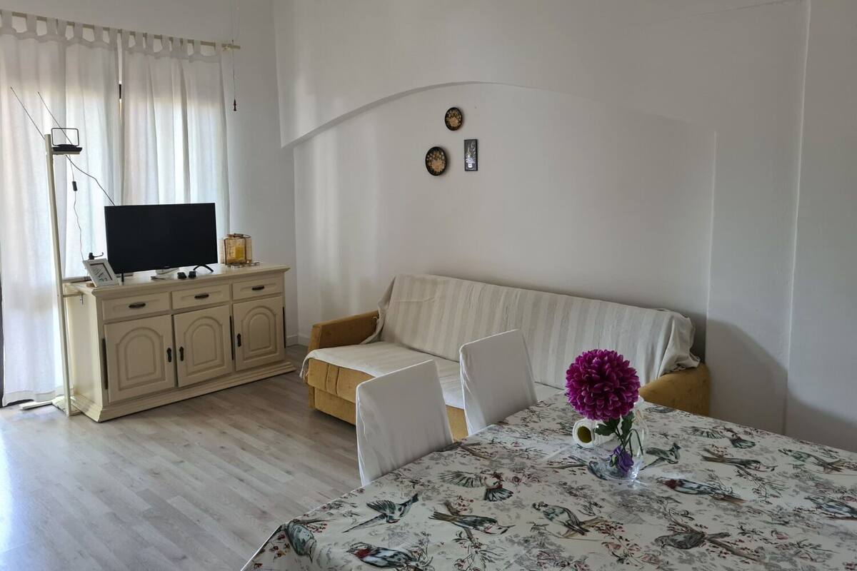 A living room with a sofa, a TV on a cabinet, a table with a floral tablecloth, and a vase with flowers.