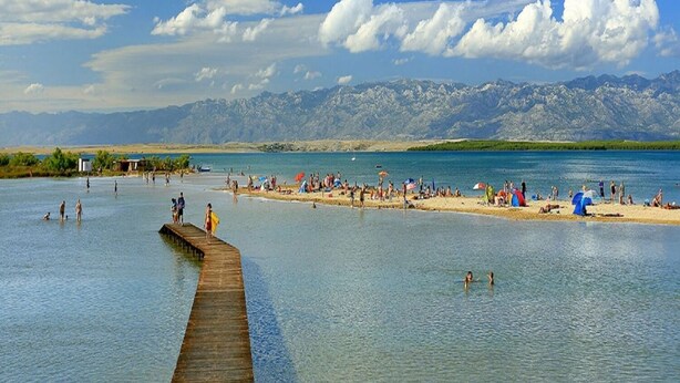 A beach with a wooden pier extending into calm waters, surrounded by mountains and clear skies. - Apartments Stosa (Nin)