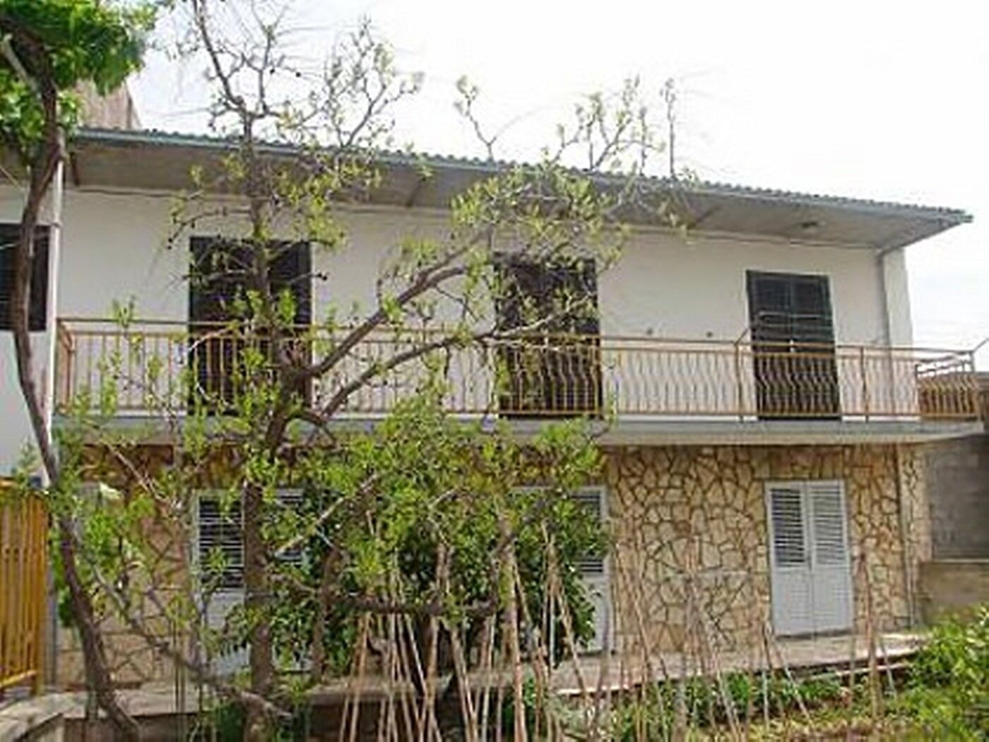 A two-story house with a balcony, stone facade, and a tree in the foreground.