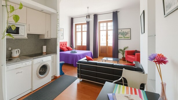 Un espace de vie compact comprenant un coin cuisine, un lit avec une literie violette, un canapé rouge et un coin repas.