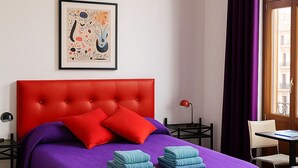 Une chambre avec un lit doté d’une tête de lit rouge à motifs capitonnés, une literie violette, un banc en bois, une œuvre d’art abstraite encadrée et une fenêtre ornée de rideaux violets.