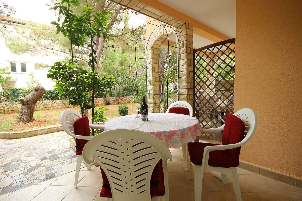 A patio with a round table and white chairs, surrounded by a stone wall and greenery.