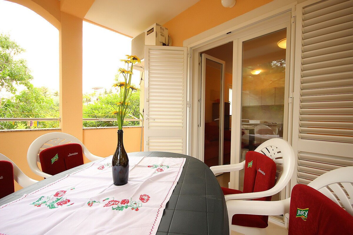 A balcony with a table set for two, white chairs, a vase with flowers, and a view of greenery.