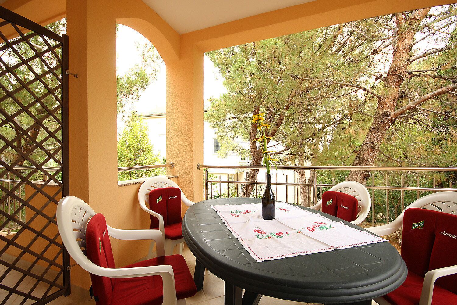 A balcony with a table and chairs, a vase with flowers, and a view of trees.