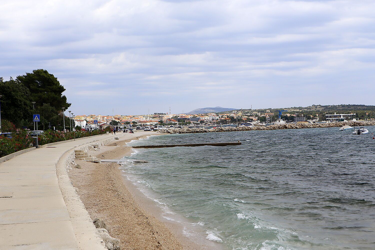 A coastal promenade with a pebble beach, a clear sky, and a distant town.