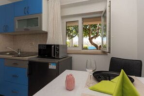 Una cocina con armarios azules, un microondas, un refrigerador pequeño, una ventana con vista a árboles y una mesa de comedor con un vaso y una servilleta verde.
