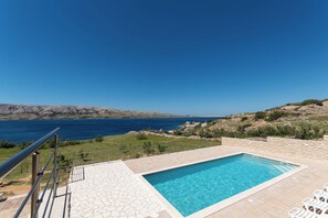 Una piscina con terraza de piedra, barandilla de metal y vistas al mar y colinas.