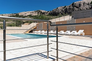 Un espace piscine doté d’un sol en pierre, de chaises longues blanches et d’un escalier menant à une terrasse.