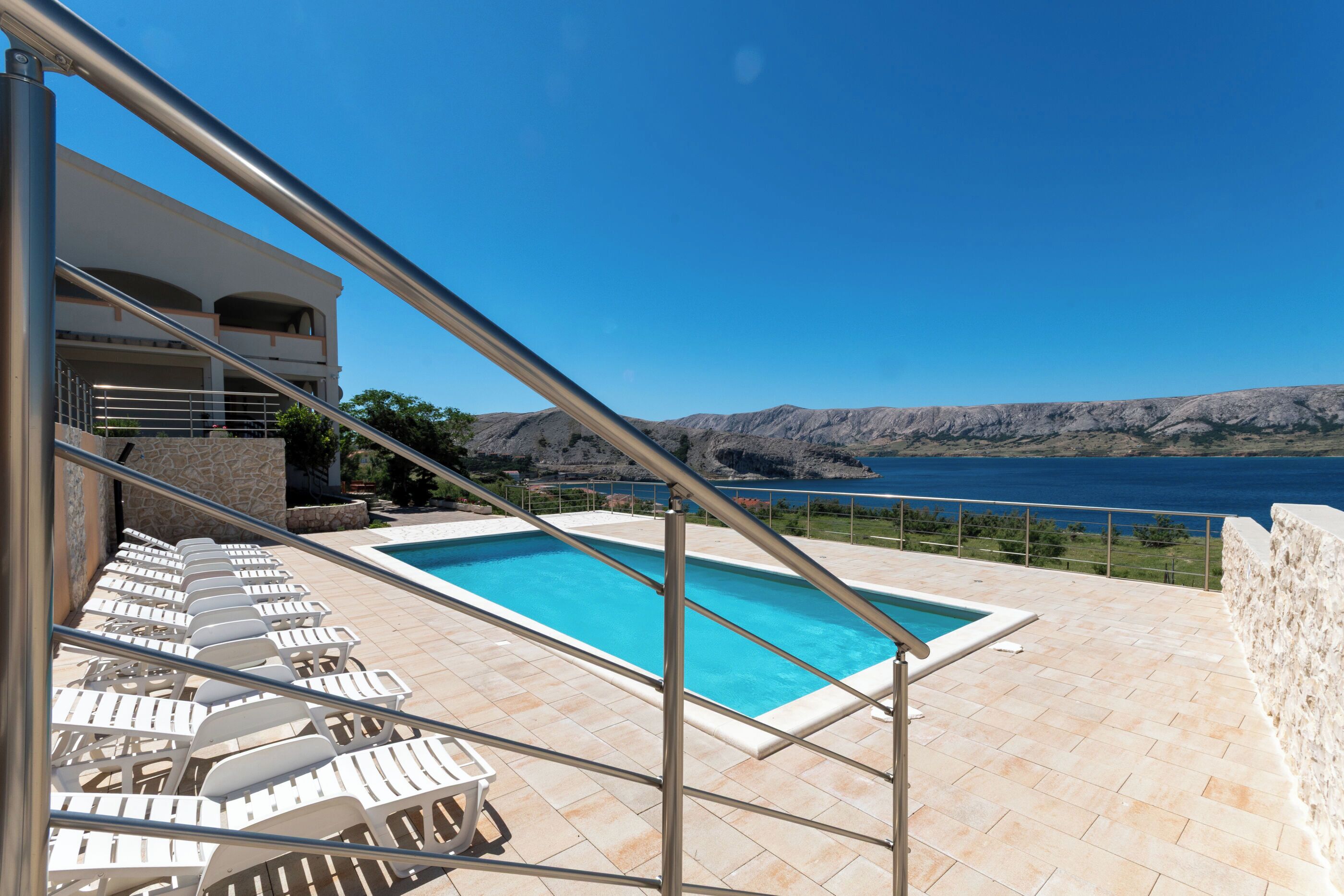 A pool with lounge chairs, a stone wall, and a view of the sea.