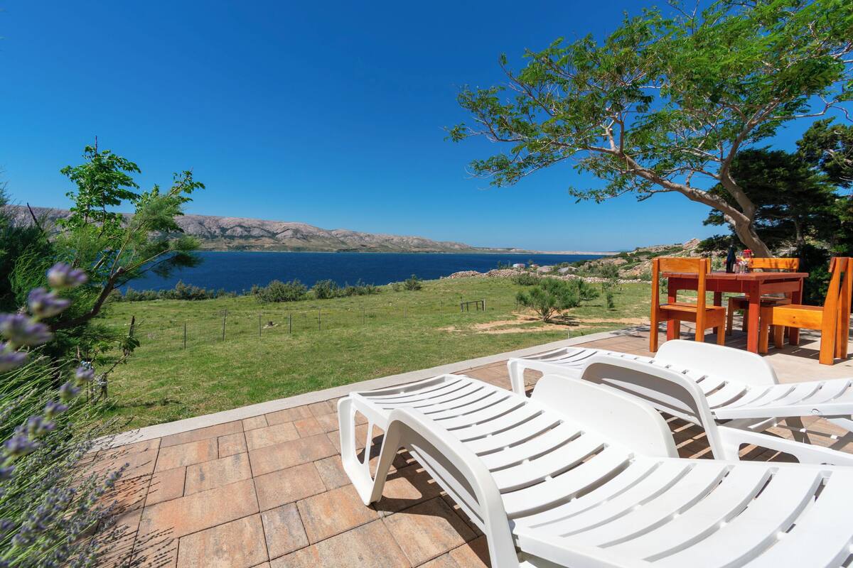 A patio with white lounge chairs, a wooden table, and a view of the sea.