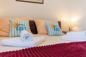 Habitación de hotel con dos camas, cada una con un almohadón con un diseño de sol y la inscripción 'Summer Vibes'. Hay toallas blancas enrolladas sobre cada cama y una manta roja en el suelo.