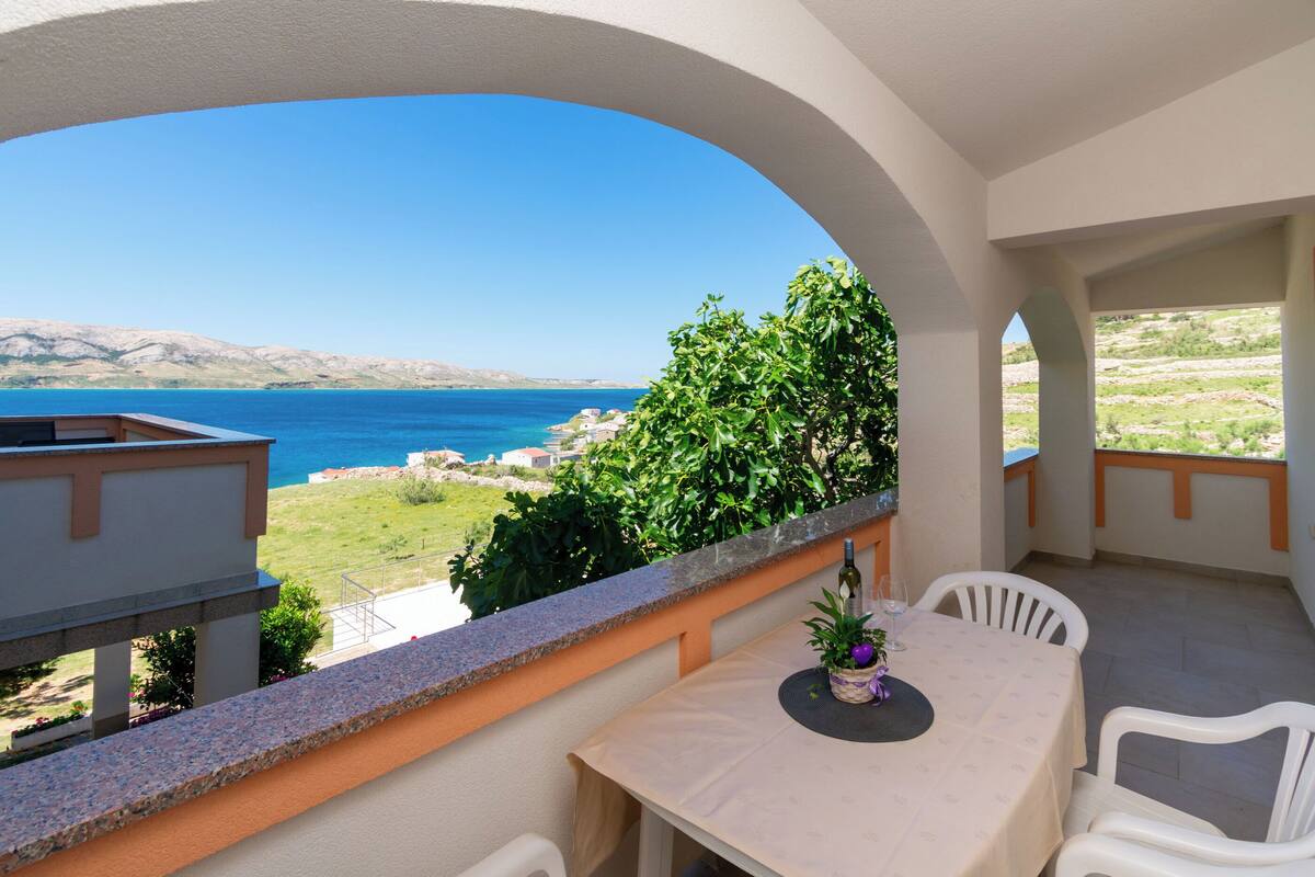 A balcony with a table and chairs, overlooking a coastal landscape with clear skies.