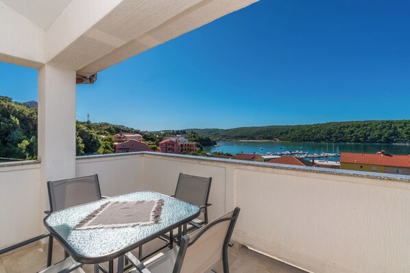 A balcony with a table and chairs, overlooking a body of water with boats and buildings in the distance. - Apartment Ren (Rab)