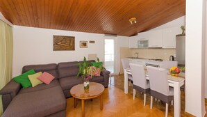 A modern living room with a wooden ceiling, a gray sofa with colorful cushions, a round coffee table with a vase of flowers, and a dining area with white chairs and a table. - Apartments Jadro (Makarska)