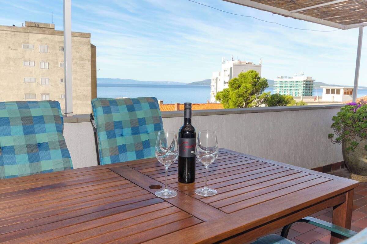 Ein Holztisch mit einer Flasche und zwei Gläsern auf einem Balkon mit Blick auf eine Skyline und das Meer.