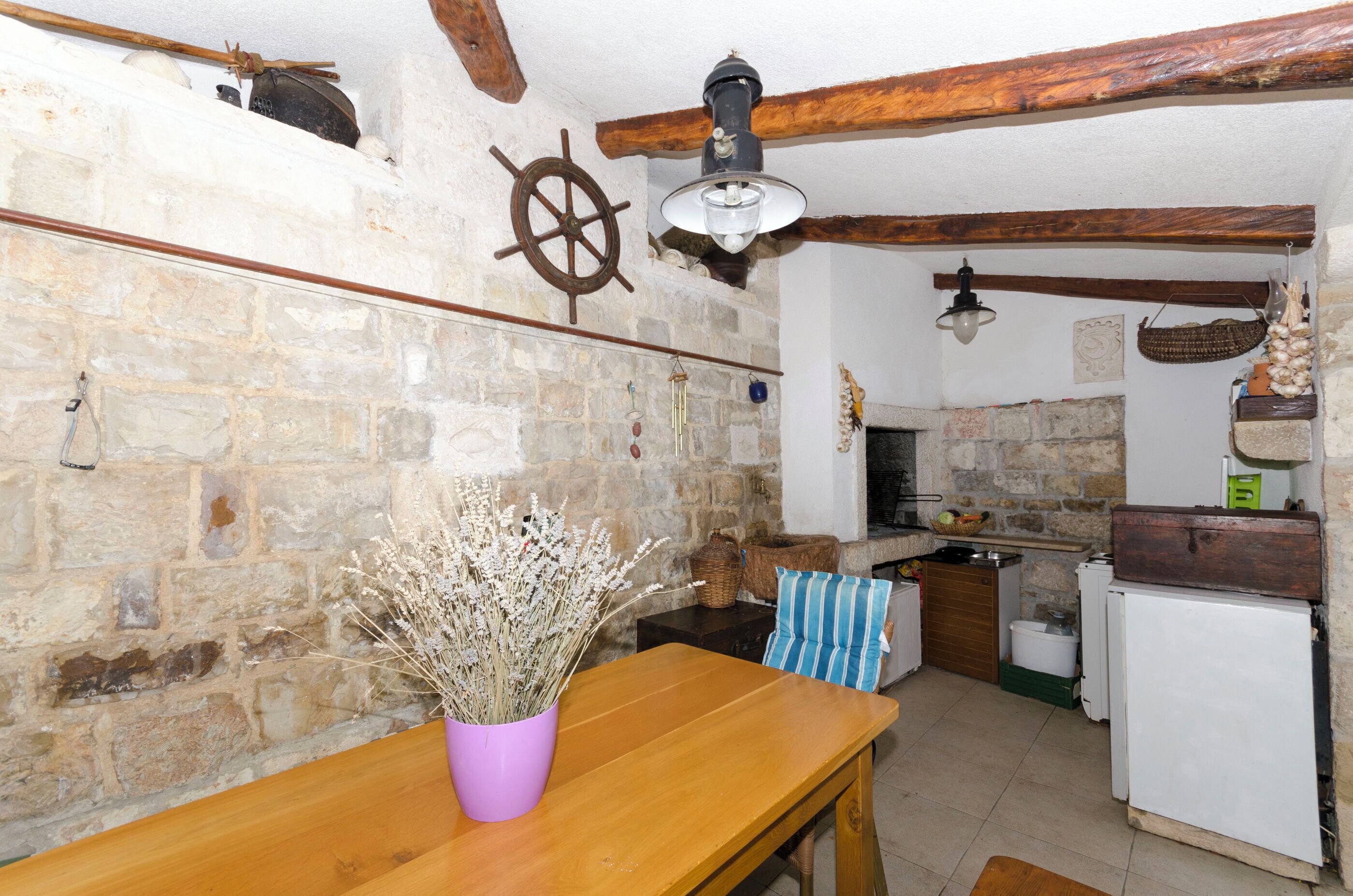 Una cocina rústica con paredes de piedra, vigas de madera, una mesa de madera con un jarrón de flores secas y una lámpara colgante.
