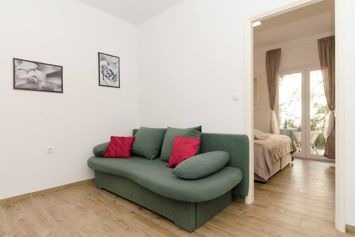 Ein grünes Sofa mit roten Kissen in einem Raum mit Holzboden und weißen Wänden. An der Wand hängen zwei gerahmte Bilder.