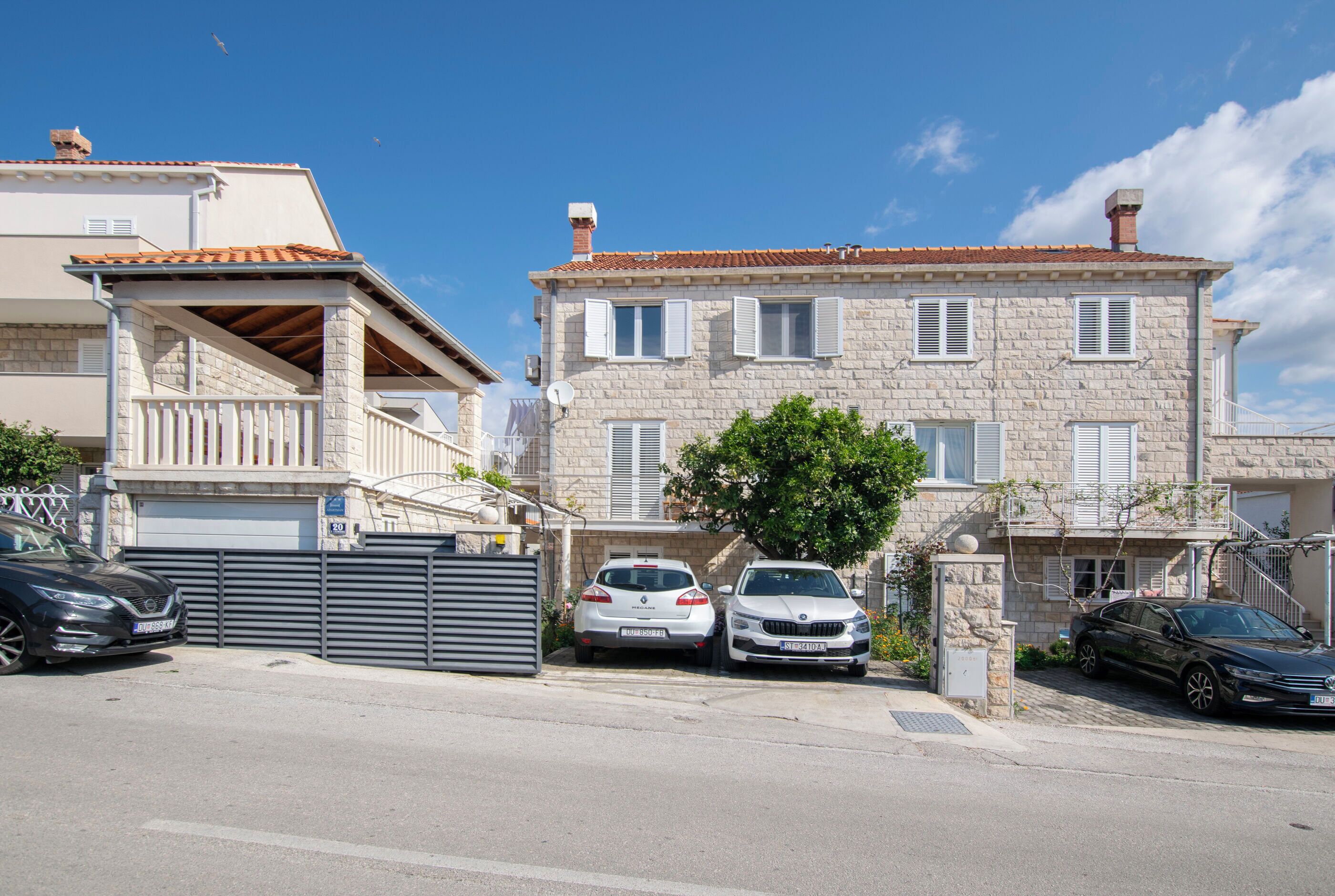 Un edificio in pietra di due piani con tetto in tegole, un vialetto recintato e auto parcheggiate.