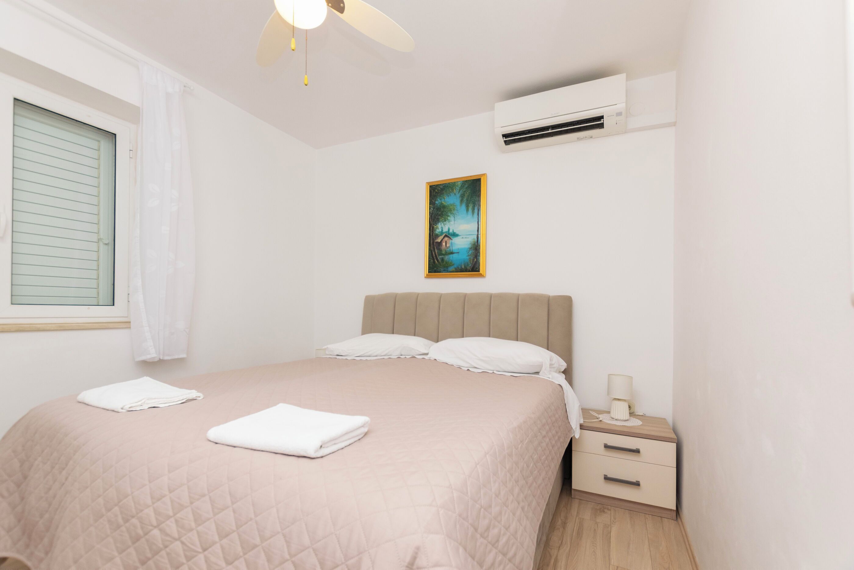 Une chambre à coucher comprenant un lit, une table de chevet, un climatiseur, une fenêtre avec des stores et un tableau accroché au mur.