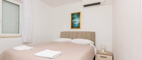 Une chambre à coucher comprenant un lit, une table de chevet, un climatiseur, une fenêtre avec des stores et un tableau accroché au mur.