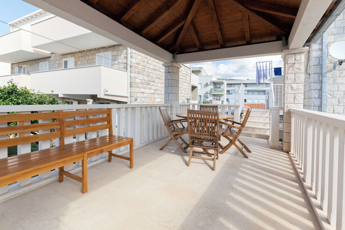 A covered outdoor seating area with a wooden bench and table, stone walls, and a view of neighboring buildings.