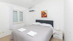Een slaapkamer met een bed, nachtkastje, raam met jaloezieën en een schilderij aan de muur.