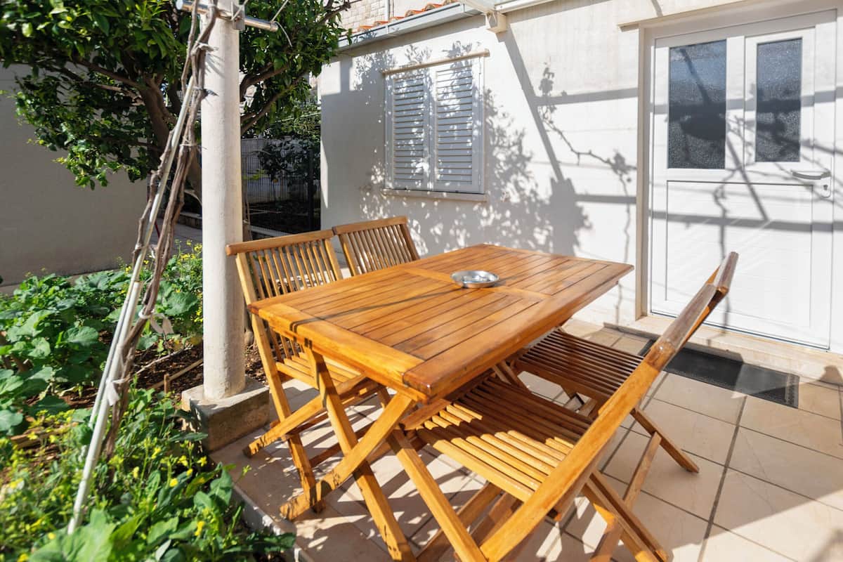 An outdoor wooden table and chairs set up on a tiled patio with a white wall and window in the background.