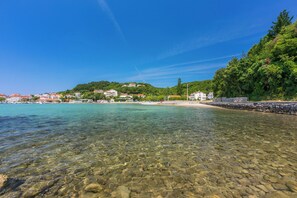 A coastal area with clear water, a sandy beach, and buildings in the background. - Apartments Ivan (Rab)