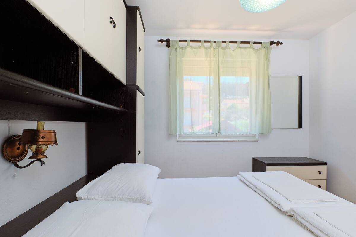 A bedroom with a bed, a bedside table, a lamp, a window with a curtain, and a mirror on the wall.