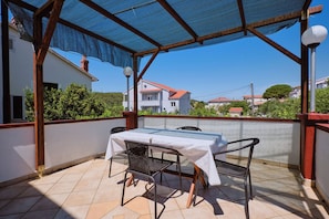 A covered patio with a table and chairs, a view of houses in the background, and a clear sky. - Apartments Ivan (Rab)