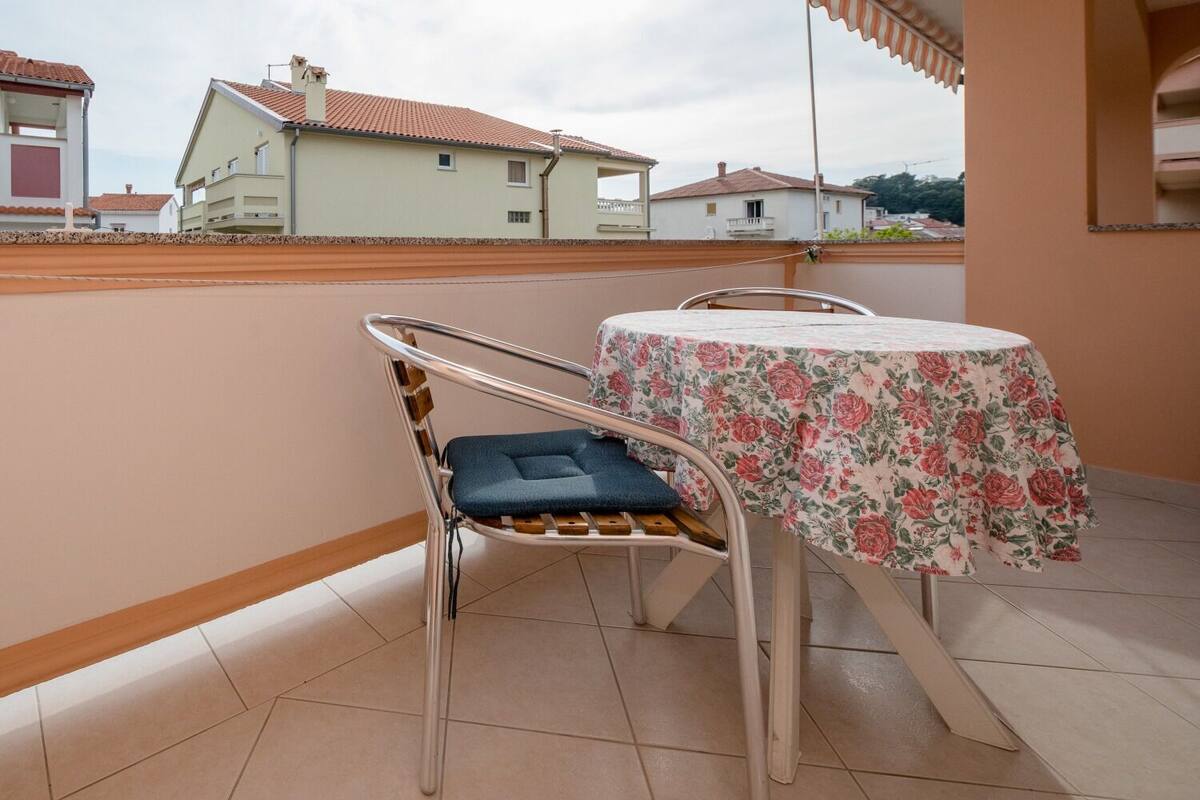Un balcón con una mesa y una silla, piso de baldosas y vista a casas al fondo.