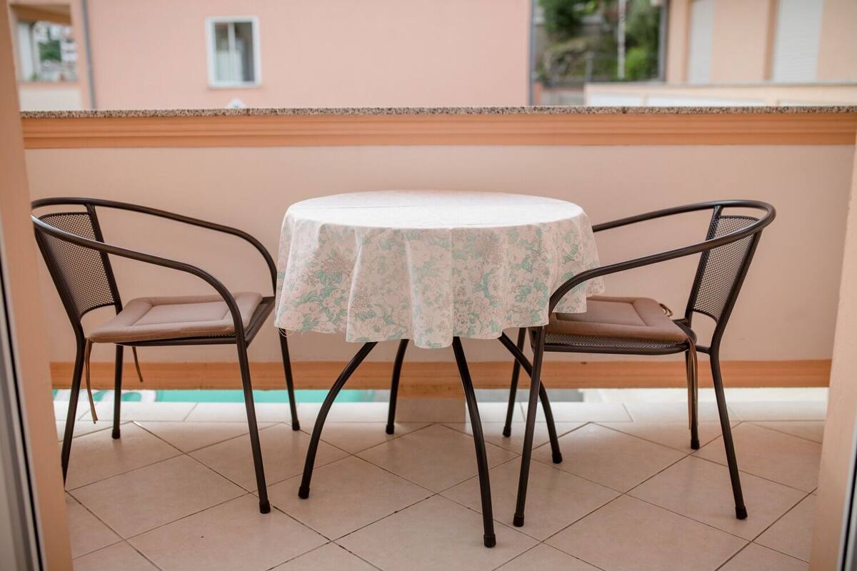 Una mesa redonda con mantel floreado, dos sillas con respaldo de malla y vista a una piscina a través de un ventanal.